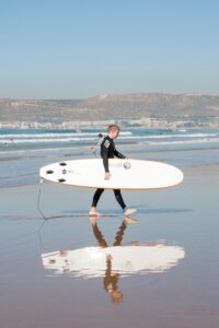 A surfer walks along Agadir beach carrying a surfboard, reflecting in wet sand.